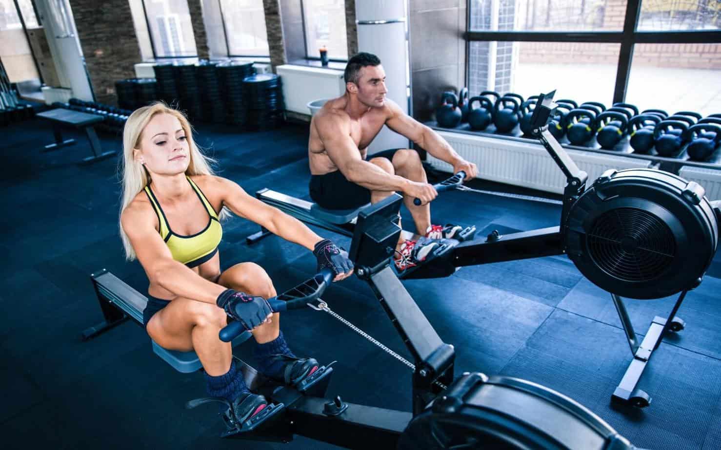 muscular man and sporty woman doing rowing hiit workouts on training simulator in crossfit gym